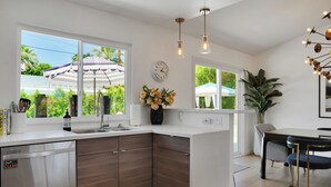 A modern kitchen with a built-in dishwasher, wooden cabinets, a large window with an umbrella, and a dining area with striped chairs. - Stylish Desert Oasis - Private Pool & Spa, 3BR/2BA Luxury Stay close to Downtown (Palm Springs)