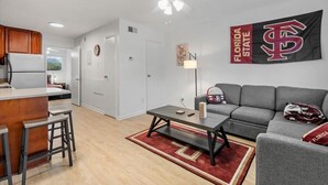 Una sala de estar moderna con un sofá gris, una mesa de centro y una zona de cocina con refrigerador y armarios de madera.