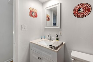 Une salle de bain avec un lavabo blanc, un miroir et des toilettes. Deux éléments décoratifs ornent les murs : l’un représente la région des Seminoles et l’autre, l’université d’État de Floride.