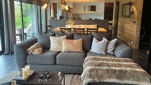 A modern living room with a grey sofa, a coffee table, and a fur throw. There is a dining area with a wooden table and chairs in the background. - Riverside Cottage (Skorradalshreppur)