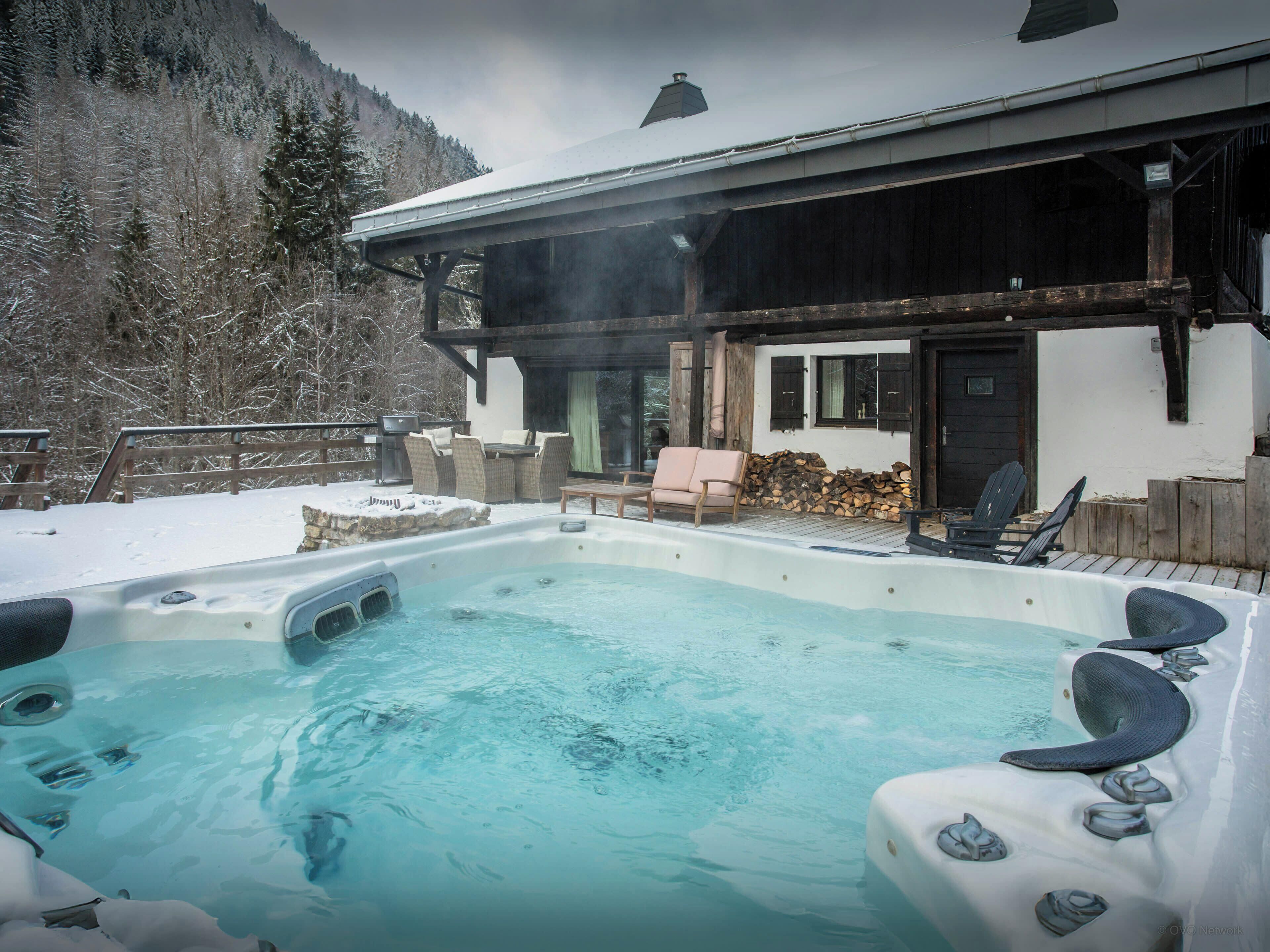 Un bain à remous installé à l’extérieur, dans un décor enneigé, avec une terrasse en bois, des chaises et un bâtiment en arrière-plan.