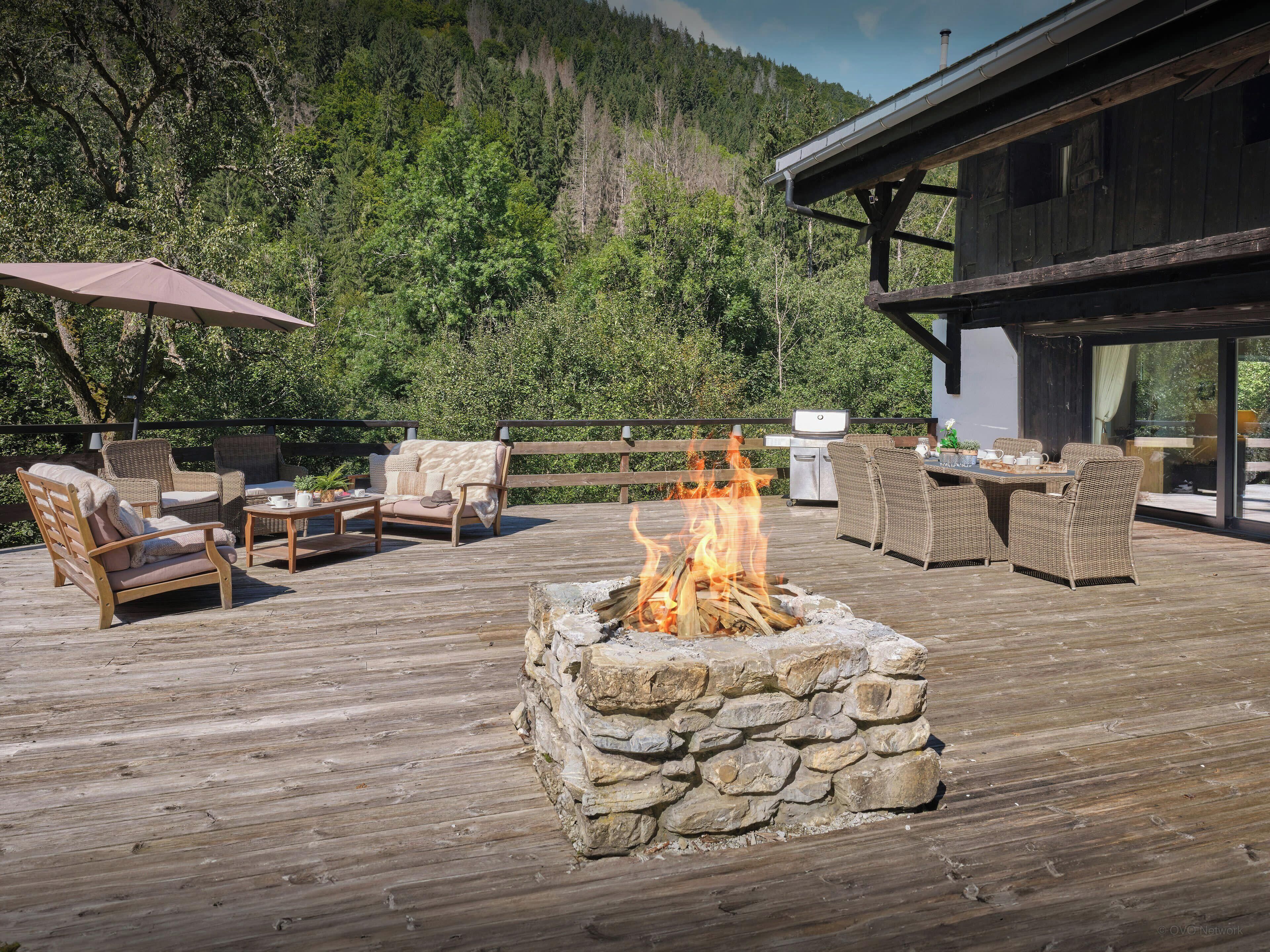 A wooden deck with outdoor furniture, a stone fire pit, and a view of a forested mountain.