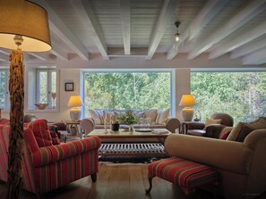 Una sala de estar acogedora con techo de madera, ventanas amplias con vistas a zonas verdes, sofás a rayas y una mesa de centro.