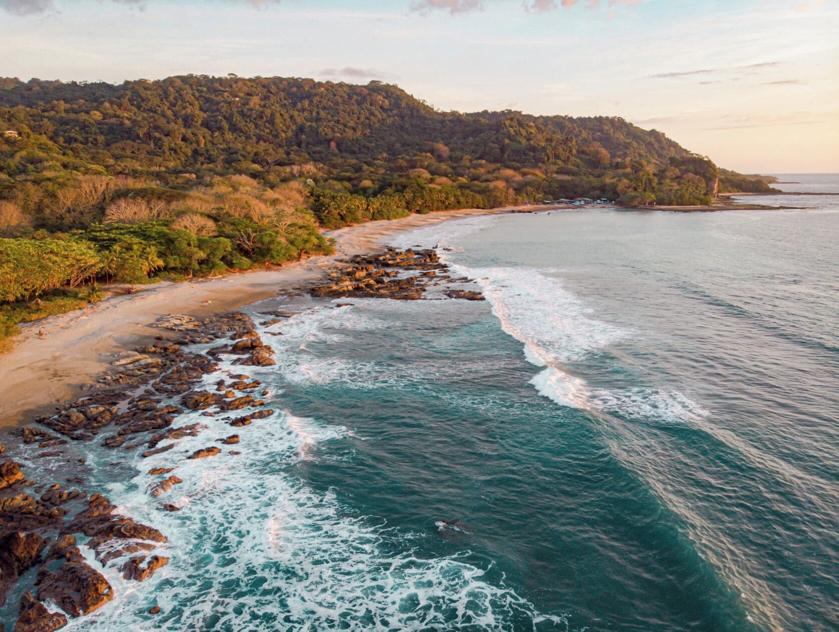 A coastal landscape with a beach, rocky shoreline, and forested hills.