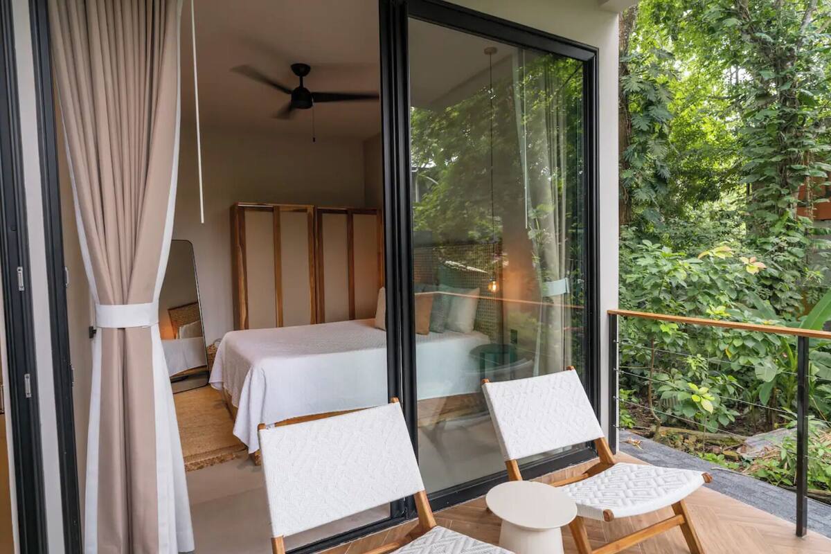 A bedroom with a bed, a ceiling fan, and a glass door leading to a balcony with chairs and a small table.