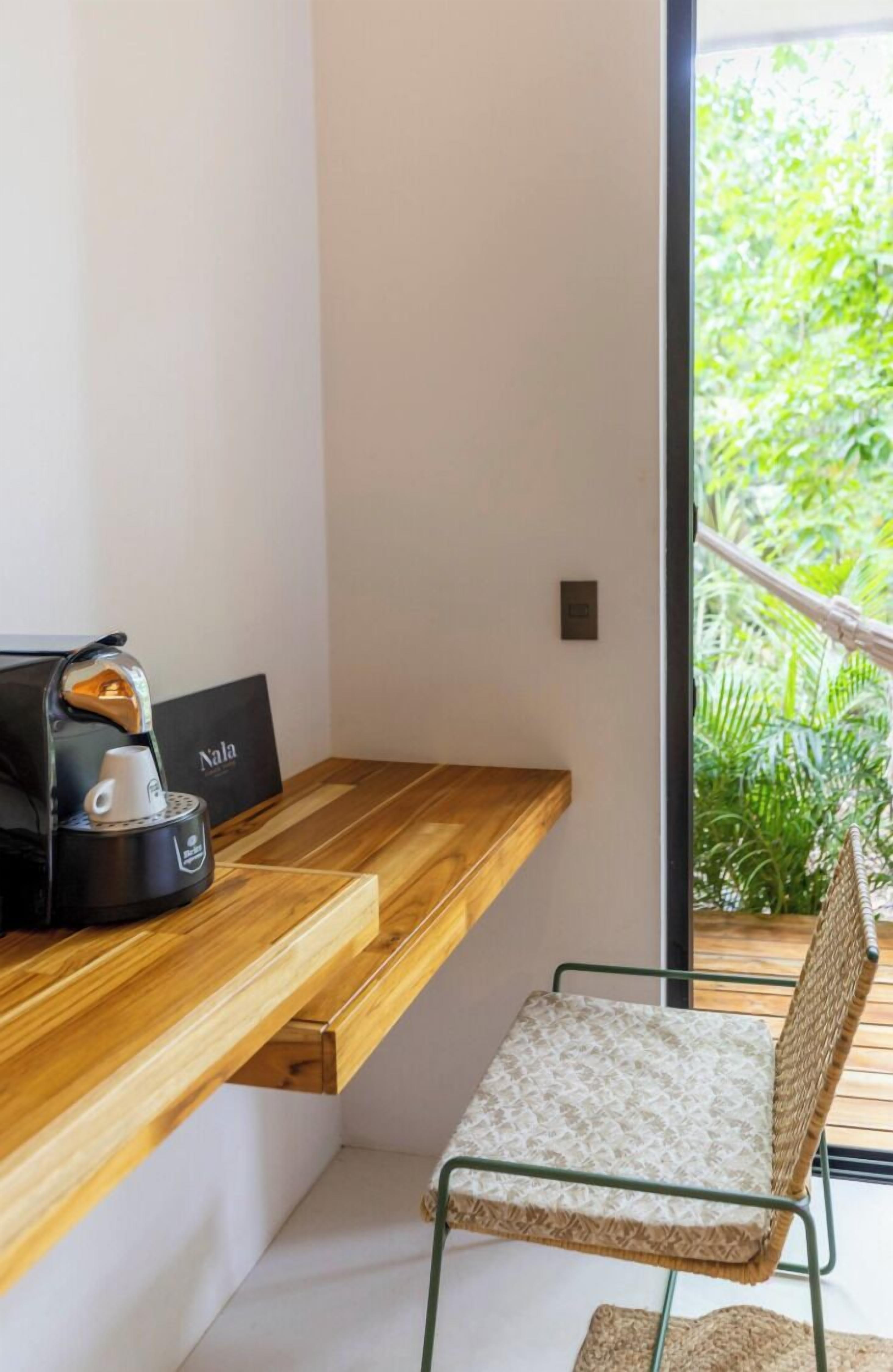 Habitación moderna con escritorio de madera, cafetera Nala, silla de mimbre y vista a zonas verdes a través de un ventanal amplio.