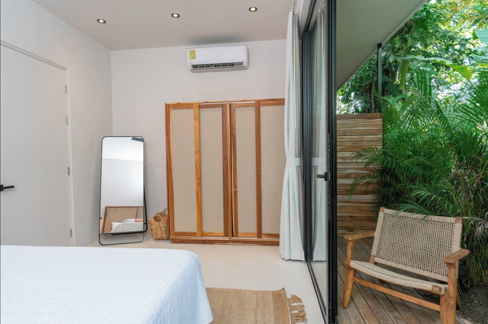 Quarto com cama, espelho, guarda-roupa e vista para um jardim exuberante através de uma porta de correr.