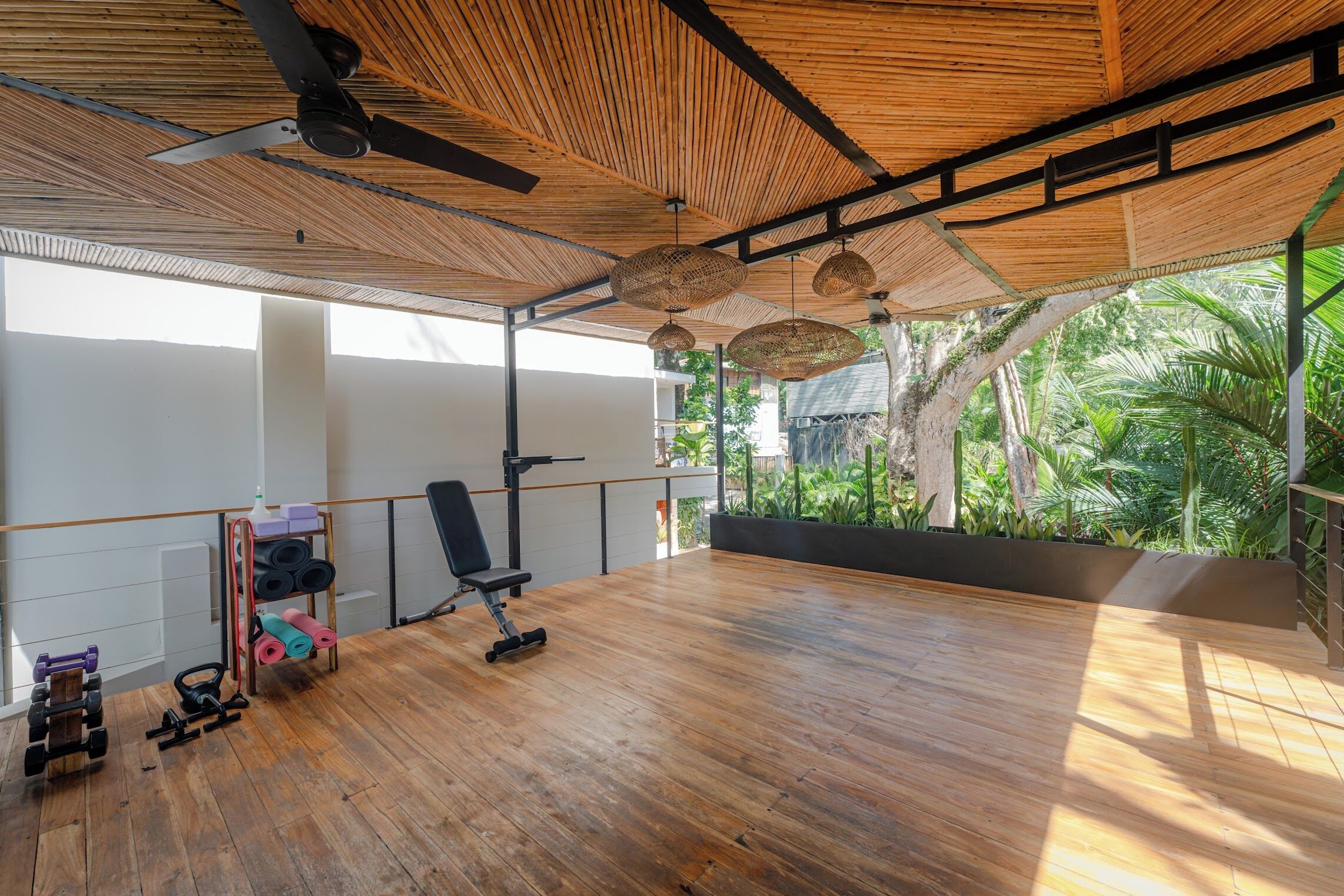 Amplia habitación con piso de madera, ventiladores de techo, equipo de ejercicio y vista a la vegetación exuberante.