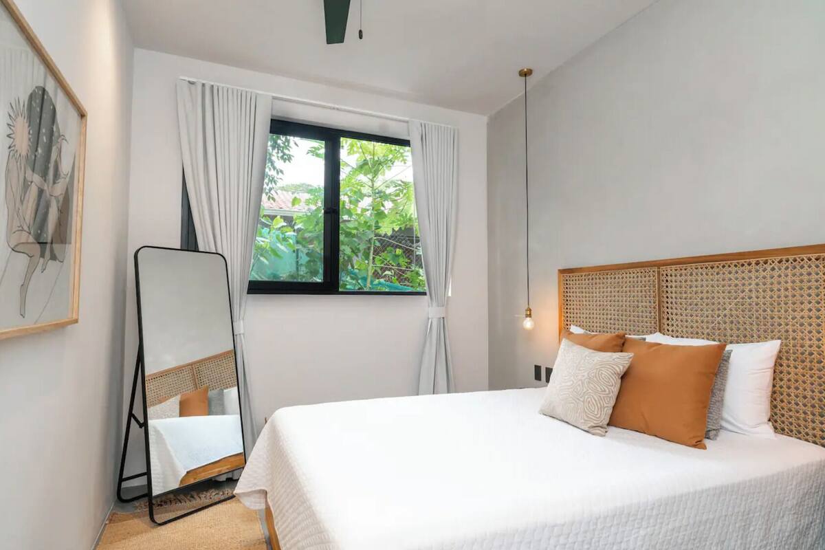 A neatly arranged bedroom with a large bed, a standing mirror, and a window with curtains.