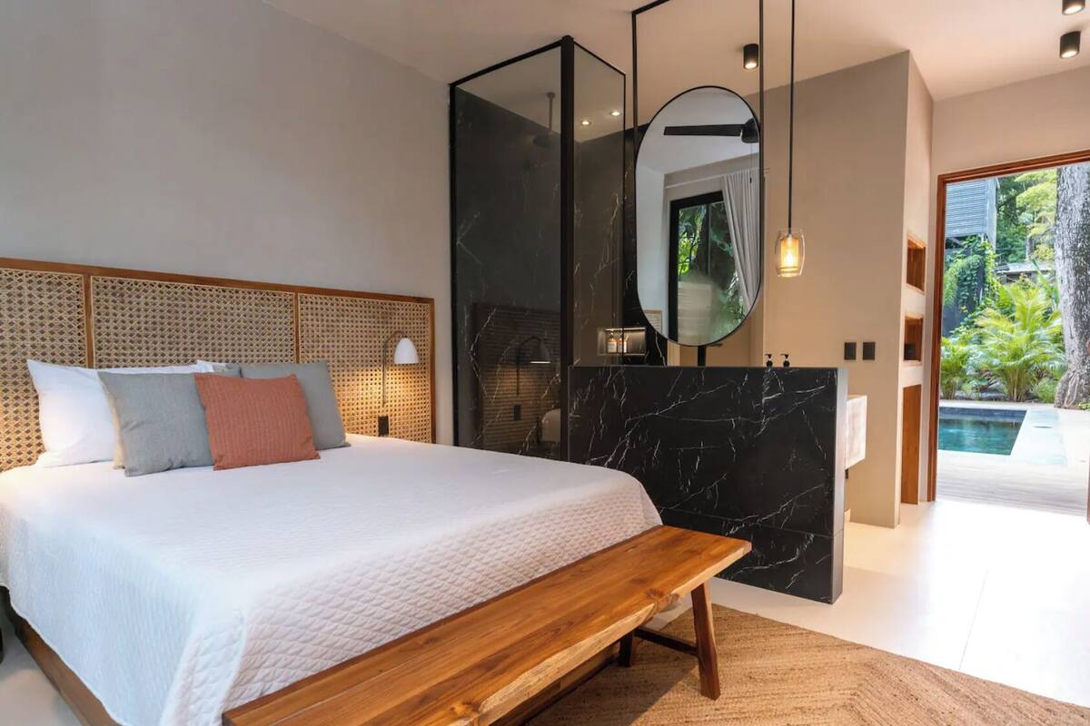 A modern bedroom with a large bed, a wooden bench, a bathroom with a marble vanity, and a view of a tropical garden.