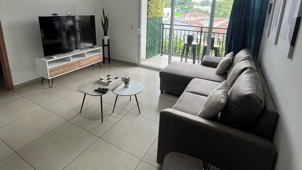 Una sala de estar moderna con un televisor de pantalla plana, un sofá, una mesa de centro y un balcón con vistas al vecindario.