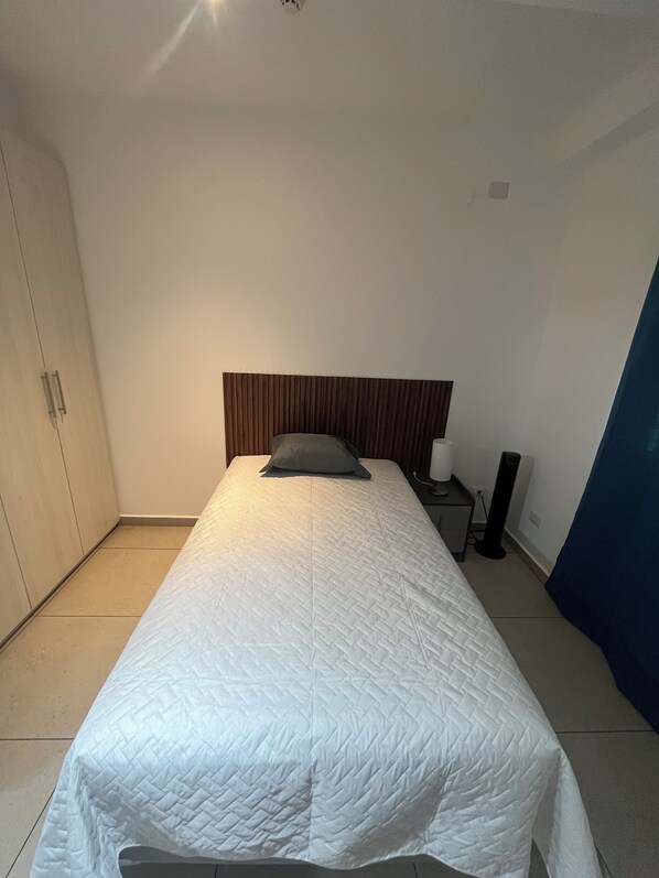 Una cama bien hecha con una colcha acolchada blanca y una sola almohada, un cabecero de madera, una mesita de noche con una lámpara y un armario.