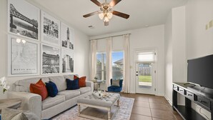 A living room with a grey sofa, blue chairs, a coffee table, and a TV on a black stand. There are framed pictures on the wall and a ceiling fan.