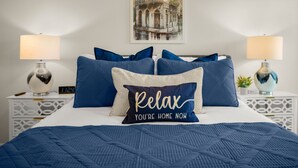 A neatly made bed with a blue quilt, a pillow with "Relax You're Home Now" text, and a digital clock on a bedside table.