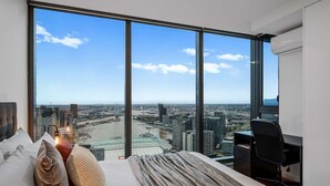 A bedroom with a large window offering a city view, a bed with pillows, and a desk with a chair.