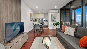 A modern living room with a flat-screen TV, a sofa with cushions, a dining area with chairs, and a kitchen in the background.