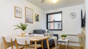 A modern living room with a dining area, a grey sofa, a small side table with a plant, and a large window with a view of a brick building.