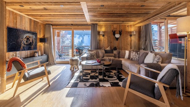 A cozy cabin interior with a wooden ceiling, a large window with a snowy view, a mounted deer head, and a comfortable seating area with a sofa and chairs. - Appartement Élégance Mont Blanc - Welkeys (Combloux)