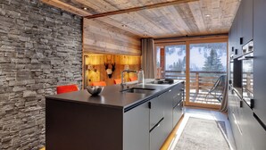A modern kitchen with a stone wall, wooden ceiling, and a large island. - Appartement Élégance Mont Blanc - Welkeys (Combloux)