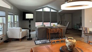 A spacious living room with a high ceiling, large windows, a modern chandelier, a sofa, a coffee table, and a television mounted on the wall.