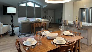 A modern kitchen with a stainless steel refrigerator, a dining area with a wooden table set for four, and a living room with a sofa and a TV.
