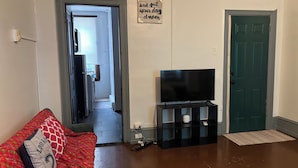 A living room with a TV on a stand, a bed with a red and white patterned blanket, a door, and a hallway leading to a kitchen. - Temple Hosp 2nd floor (Philadelphia)