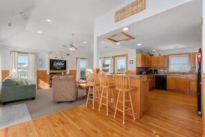 A spacious living area with a sofa, a coffee table, and a TV. There is a kitchen with wooden cabinets and a bar area with stools.