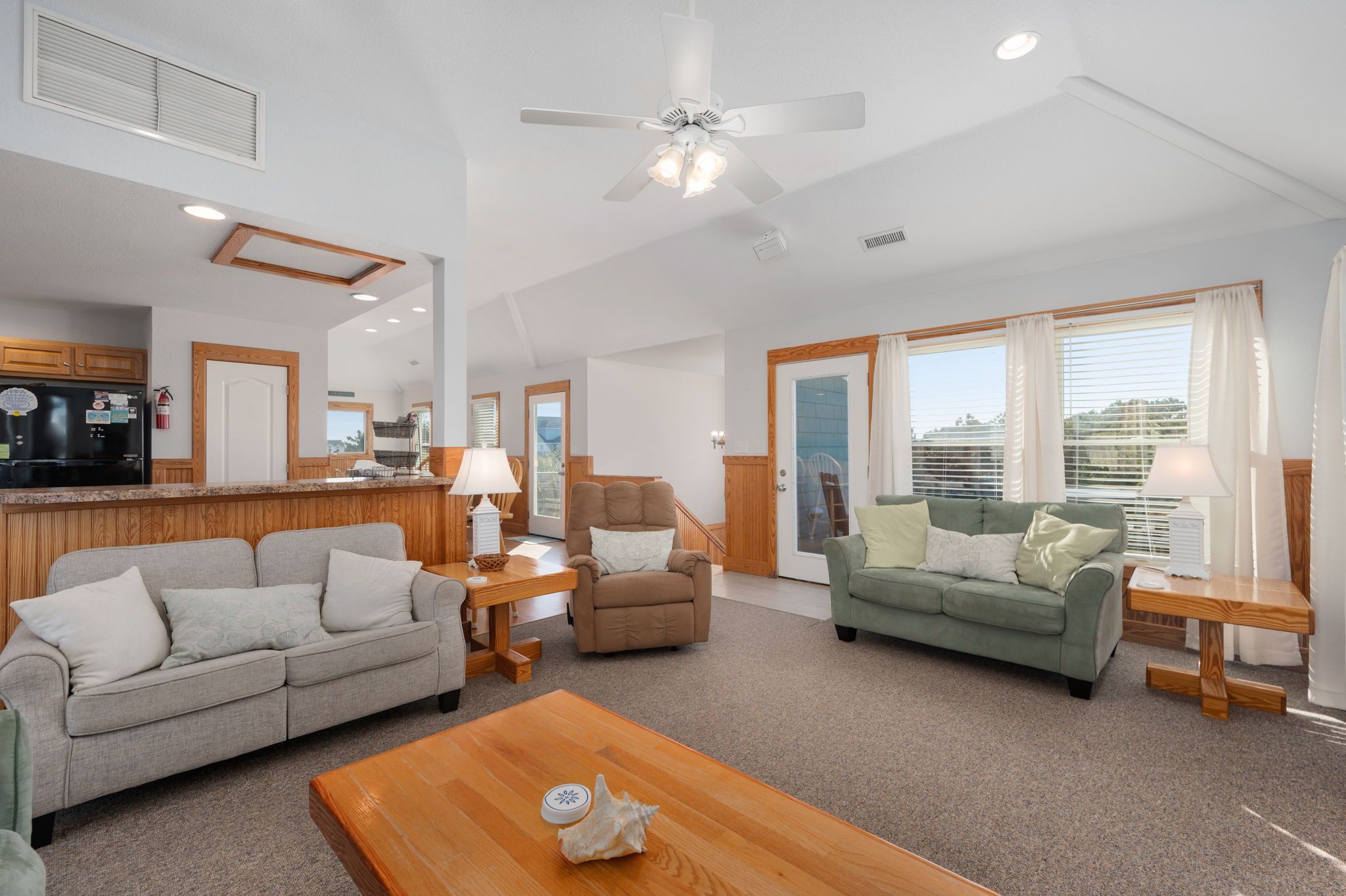 A spacious living room with a wooden coffee table, a beige sofa, a green sofa, and a recliner. There is a kitchen area with wooden cabinets and a refrigerator.
