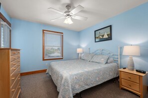 A bedroom with a bed, a dresser, a nightstand, a ceiling fan, a window with blinds, and a painting on the wall. - Beachnuts Too: Pet Friendly, Oceanside, Private Pool and Hot Tub (Corolla)