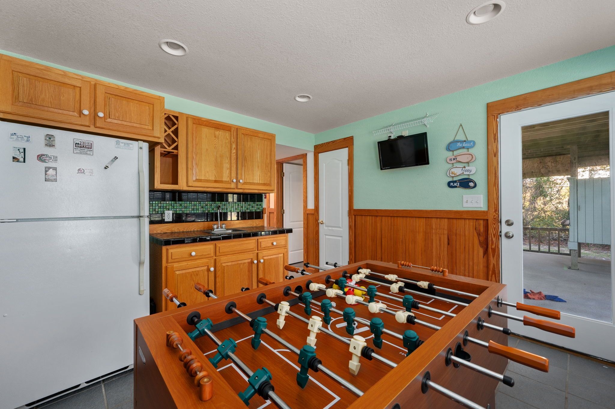 A kitchen with wooden cabinets, a refrigerator, a foosball table, and a door leading to an outdoor area.