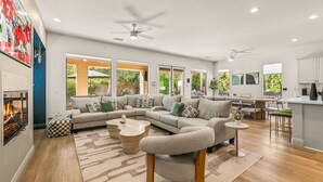 A spacious living room with a large sectional sofa, a fireplace, and a dining area with a kitchen in the background. - Rare Gem: Pool, Spa, Game Room, Fire Table! (Indio)
