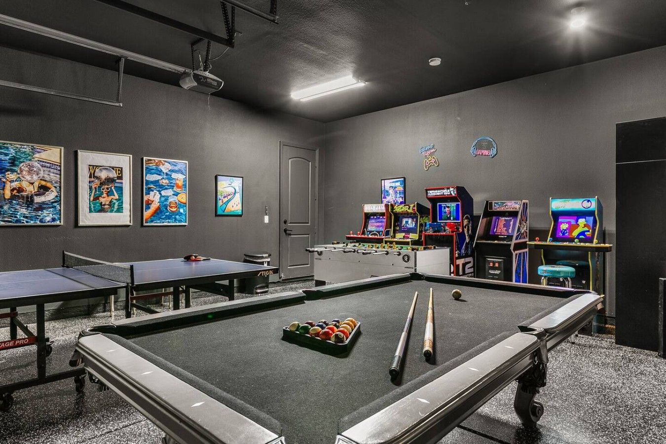 A pool table with billiard balls, a ping pong table, and arcade machines in a room with grey walls and ceiling.