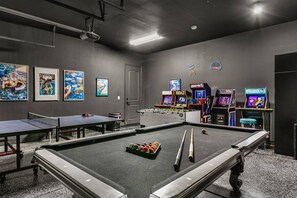 Una mesa de billar con bolas, una mesa de ping pong y máquinas de videojuegos en una sala con paredes y techo grises.