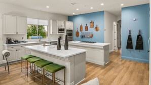 A modern kitchen with white cabinetry, a central island with stools, and a blue accent wall with hanging kitchen tools. - Rare Gem: Pool, Spa, Game Room, Fire Table! (Indio)