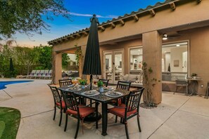 Un patio exterior con una mesa de comedor dispuesta para seis personas, rodeada de sillas y una piscina al fondo.