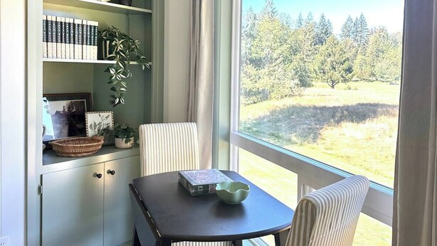 A room with a dining table and chairs, a bookshelf, and a large window with a view of trees and grass. - Charming Condo Retreat (Welches)