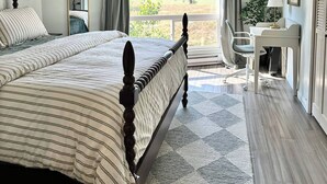 A bedroom with a large bed, a desk, and a view of a green landscape through the window. - Charming Condo Retreat (Welches)