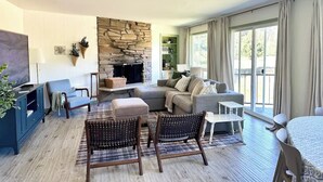 A spacious living room with a stone fireplace, a sofa set, a television, and a dining area with a table and chairs. - Charming Condo Retreat (Welches)