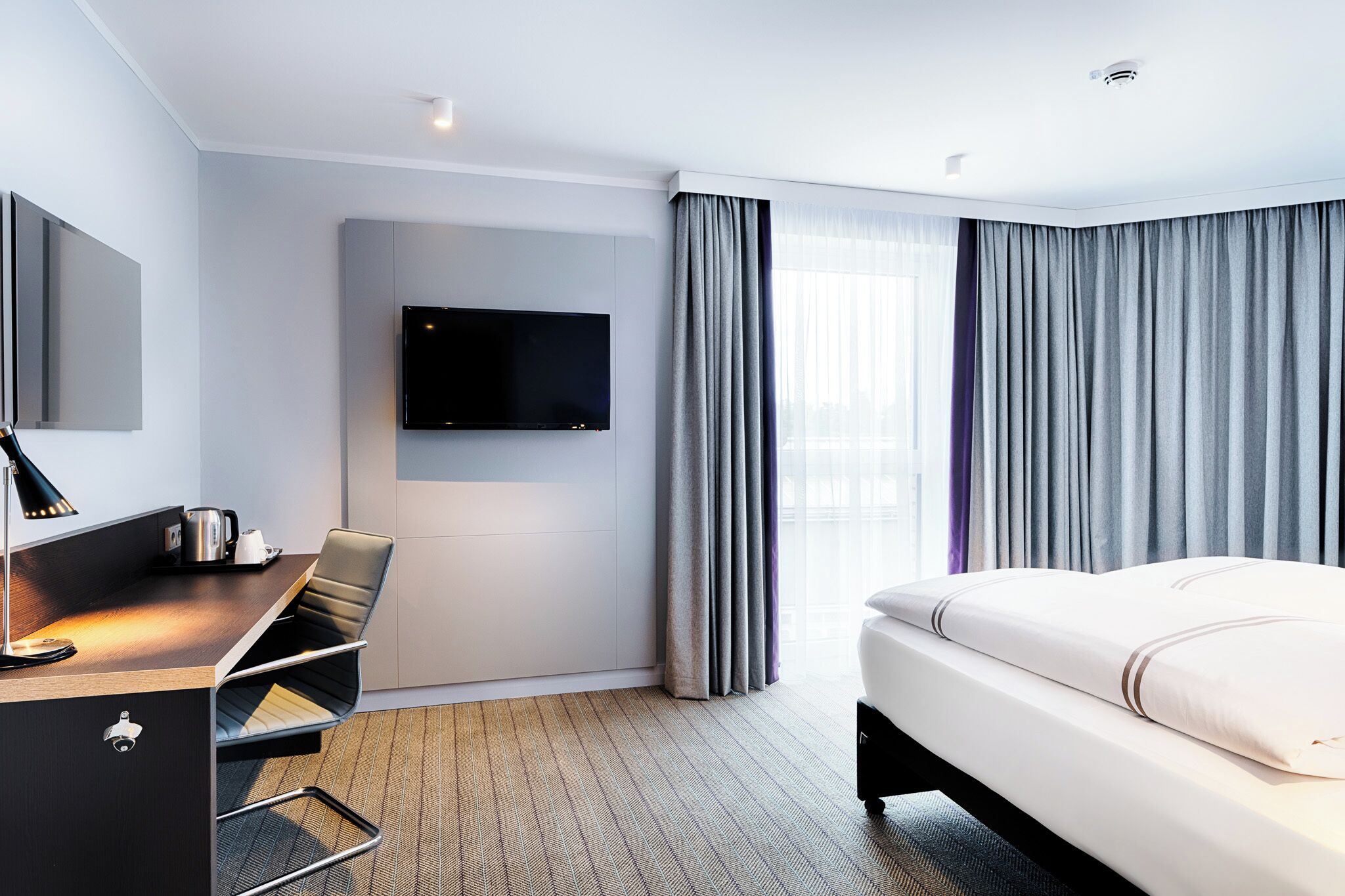 Une chambre d’hôtel moderne dotée d’un téléviseur à écran plat fixé au mur, d’un bureau avec une chaise, d’un lit avec une literie blanche et de rideaux gris.