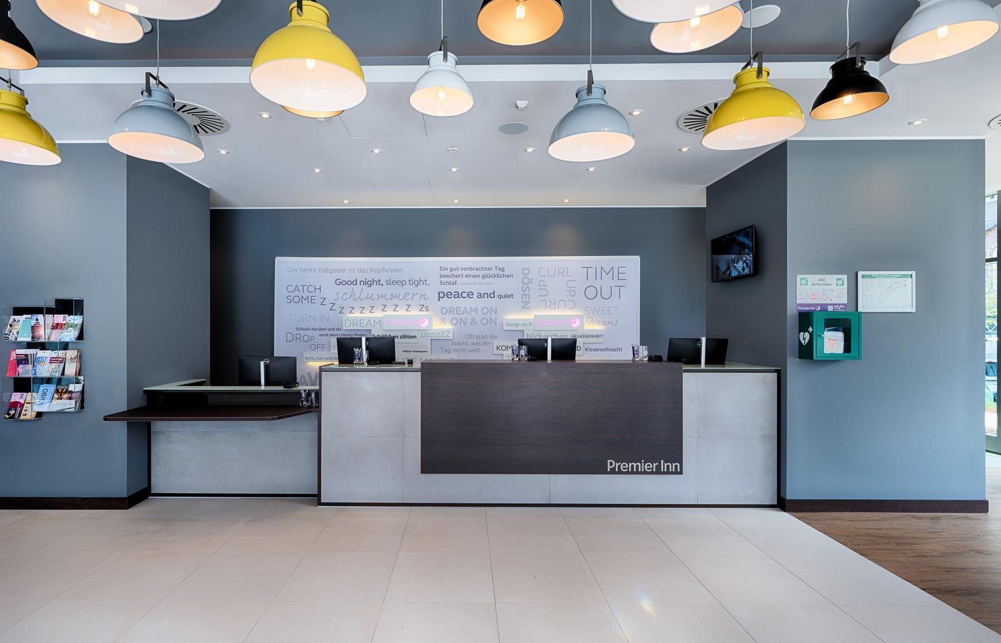 Zona de recepción de un hotel con diseño moderno, que incluye un mostrador de recepción, una gran pared con citas motivacionales y varias lámparas colgantes.