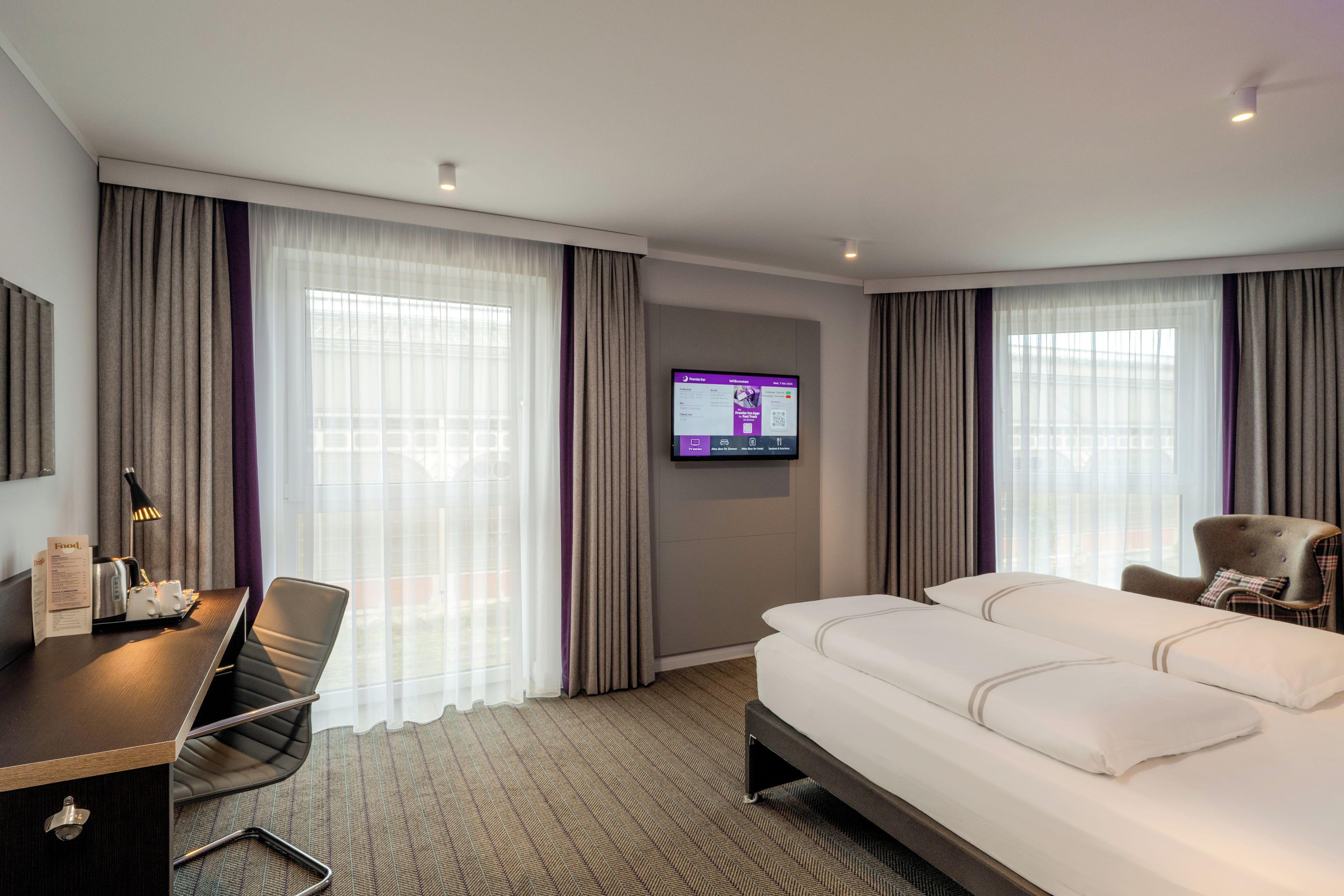 Ein Hotelzimmer mit einem Bett, einem Schreibtisch mit Stuhl, einem an der Wand befestigten Flachbildfernseher und großen Fenstern mit Vorhängen.
