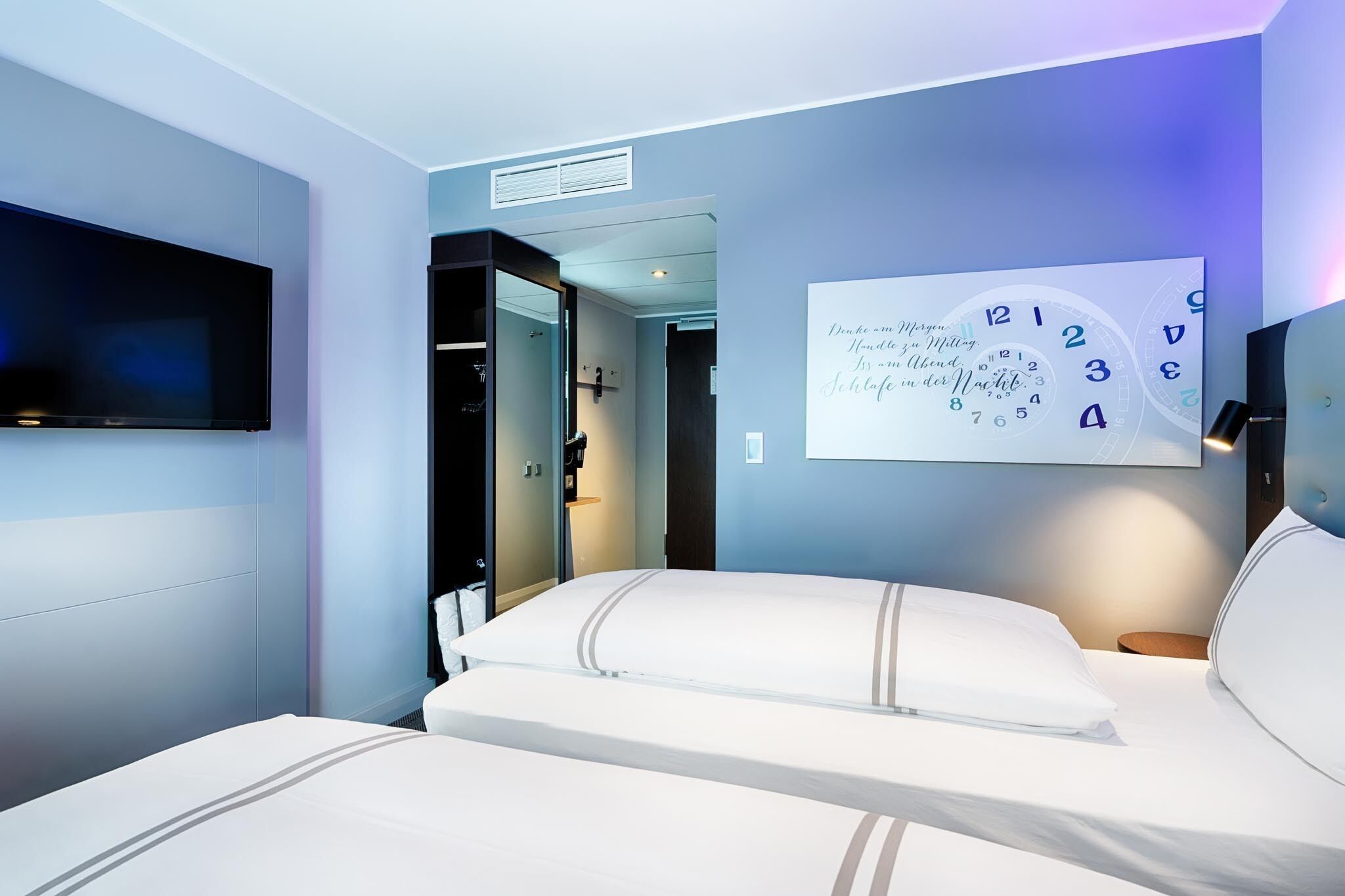Una habitación de hotel moderna con una cama grande, un televisor montado y un reloj de pared.