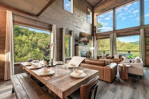 Interior de madera con una mesa de comedor dispuesta, sofás de cuero y amplias ventanas con vistas a un bosque.