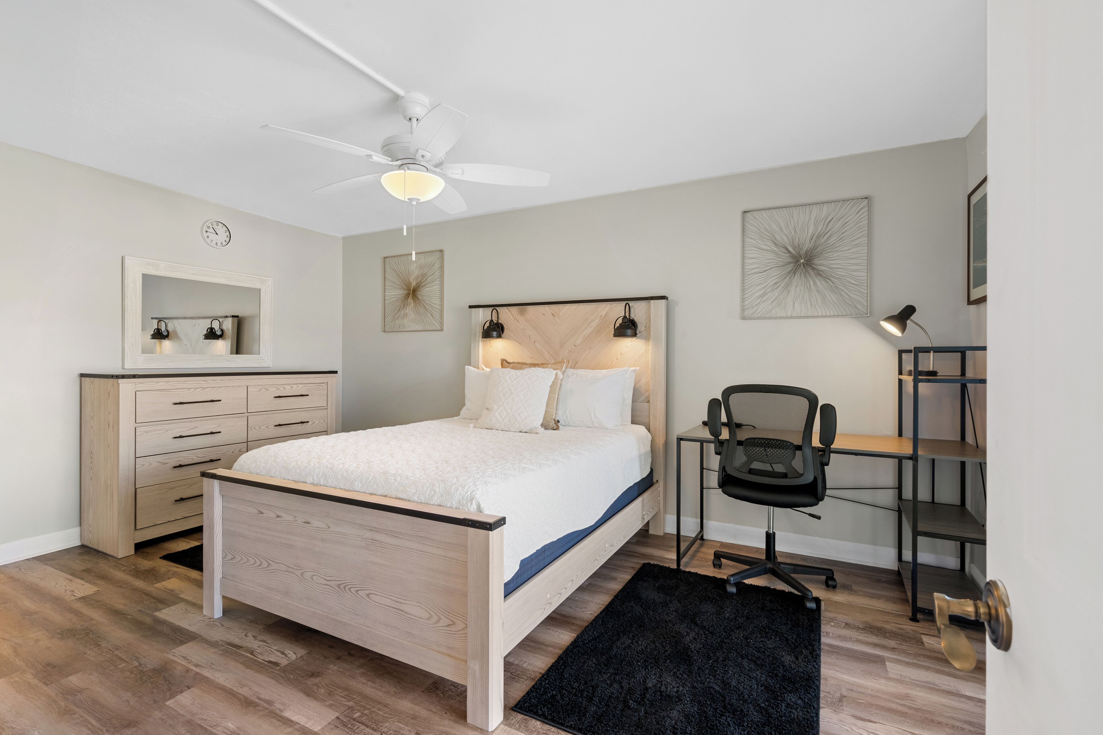 A bedroom with a bed, a desk, a chair, a ceiling fan, a clock, and a shelf.