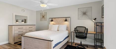 A bedroom with a bed, a desk, a chair, a ceiling fan, a clock, and a shelf.