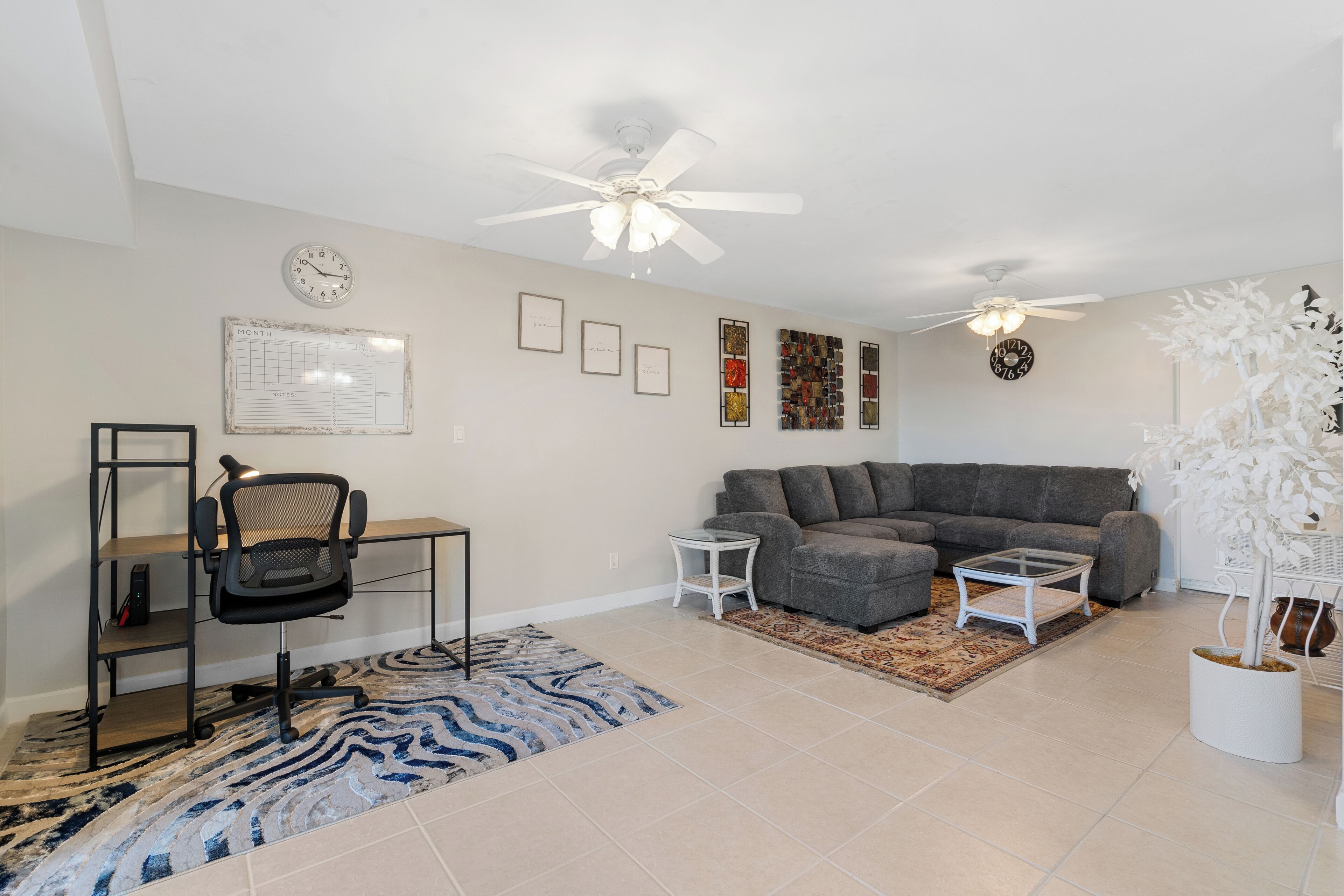 A living room with a grey sofa, a black office chair, a small white side table, a blue and white rug, and a white potted plant.
