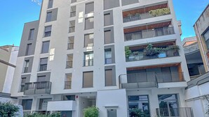 Un edificio de varios pisos con un diseño arquitectónico moderno, que cuenta con balcones con plantas, una entrada central y un área de estacionamiento con una furgoneta.