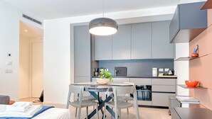 Un dormitorio moderno y funcional con cama, mesita de noche y una pequeña lámpara. Junto al dormitorio hay una kitchenette con cafetera, hervidor y una pequeña planta en maceta. El cuarto tiene un piso de madera clara y un amplio ventanal que proporciona luz natural.