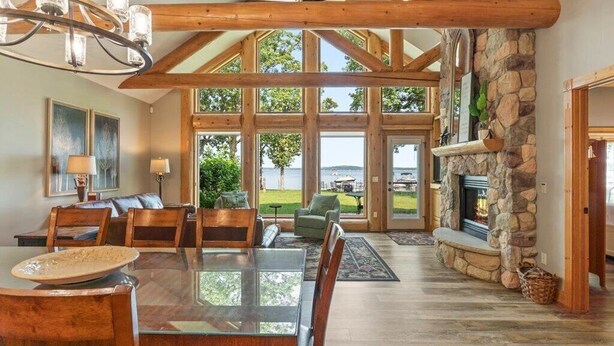 A spacious dining area with a large wooden table and chairs, a stone fireplace, and large windows offering a view of the outdoors. - Lakefront Townhome | 6BR | Man Cave + Lake View (Nisswa)