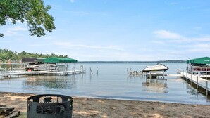 A lakeside setting with a campfire pit, docked boats, and green canopies over the water. - Lakefront Townhome | 6BR | Man Cave + Lake View (Nisswa)